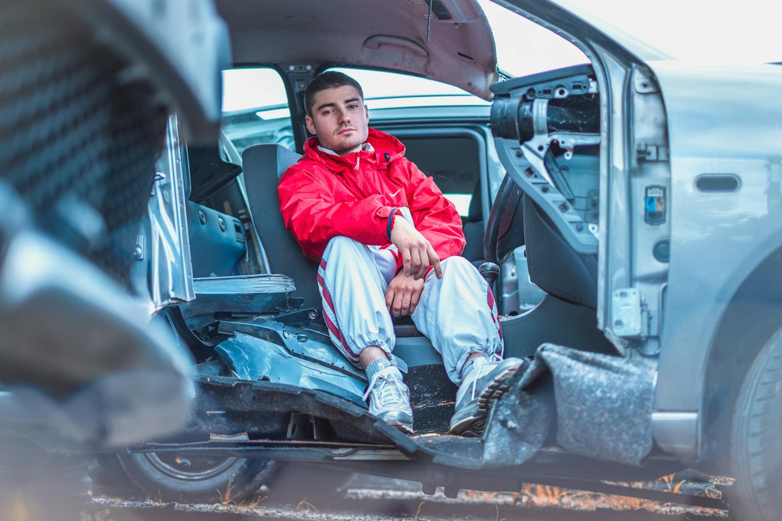 A young man in a vibrant red jacket sitting inside a damaged car, conveying themes of urban life and resilience. personal injury PIP